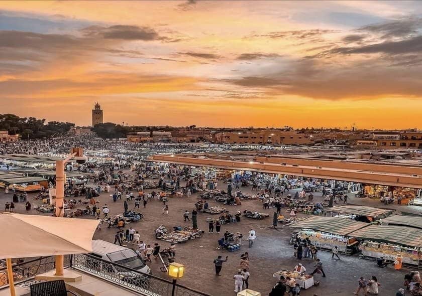 Panoramic view of Jemaa el-Fnaa Square in Marrakech, Morocco, showing bustling market stalls, crowds of people, rooftop cafés, and a golden sunset over the Medina